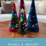 Four knitted Holiday Trees in various sizes and colors, each topped with a star and string lights, are displayed on a wooden table, with a decorated tree glowing in the background.