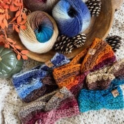 Colorful crocheted ear warmer headbands are displayed on a textured surface beside skeins of multicolored yarn, pinecones, orange leaves, and a green decorative object.