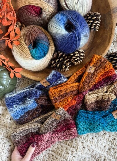 Colorful crocheted ear warmer headbands are displayed on a textured surface beside skeins of multicolored yarn, pinecones, orange leaves, and a green decorative object.