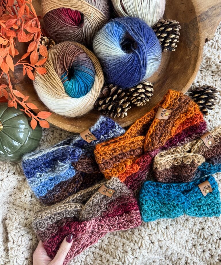 Colorful crocheted ear warmer headbands are displayed on a textured surface beside skeins of multicolored yarn, pinecones, orange leaves, and a green decorative object.