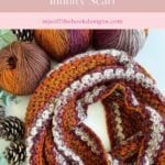 A hand holds a cozy infinity scarf in chunky crochet, featuring rich autumn colors. Around it, yarn balls and pinecones rest on a light surface, capturing the essence of seasonal warmth.