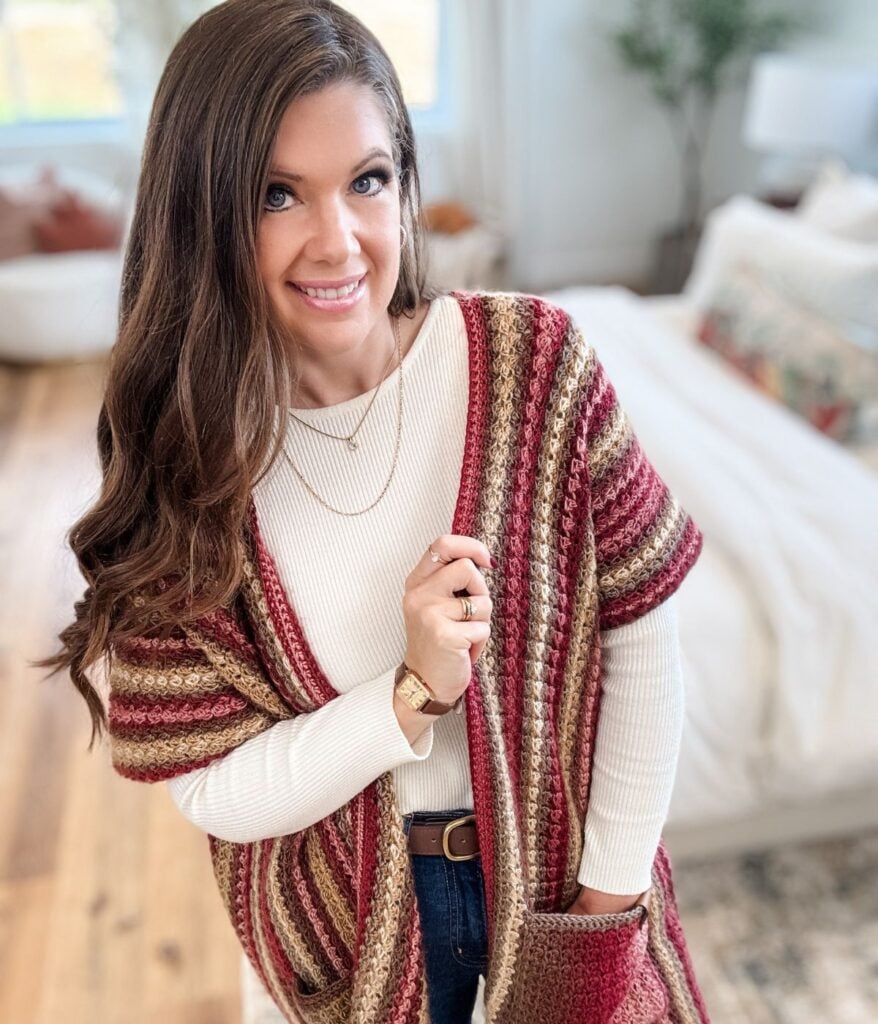 Woman with long brown hair wearing a white top and a Suzette Ruana in red, beige, and brown stripes stands in a bright, neatly arranged bedroom.
