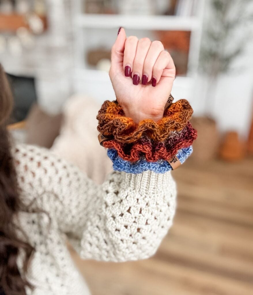A person with burgundy painted nails wears Crochet Scrunchie accessories in shades of brown, orange, and blue on their wrist, featuring the Suzette Stitch. A cream crochet sweater is also visible—a cozy look that's perfect for showcasing your next free pattern.