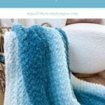 A blue and white gradient 2-day throw crochet blanket draped over the edge of a woven basket, next to a white textured chair on a wooden floor.