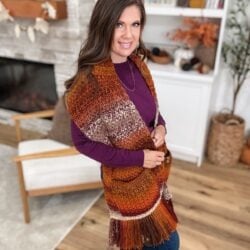 A woman stands indoors, smiling at the camera while wearing a maroon and orange knit pocket shawl with fringe. A fireplace and bookshelf serve as the cozy background.