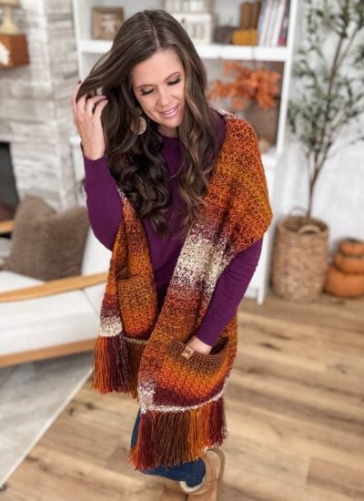 Woman wearing a purple top and a multicolored knit pocket shawl with fringe, standing in a cozy, autumn-themed living room.