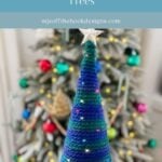 A hand holds a crocheted Holiday Tree with blue and green yarn, topped with a star, in front of a decorated Christmas tree in the background.