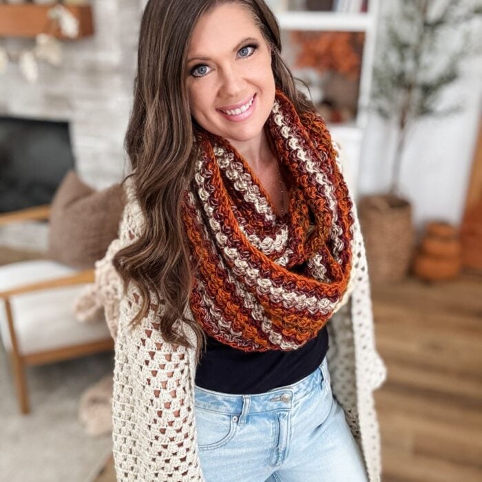 A woman with long brown hair wearing a crocheted shawl and a vibrant infinity scarf stands indoors, smiling at the camera.