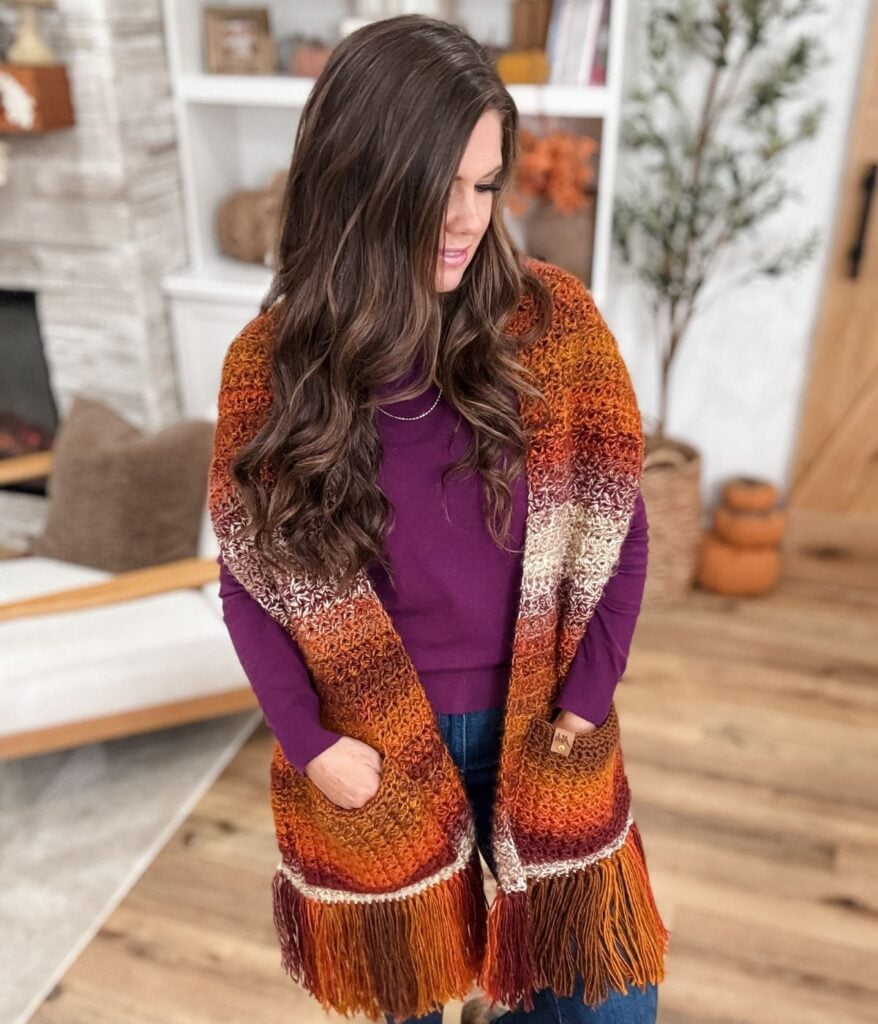 A woman with long wavy brown hair wears a purple top and a multicolored, fringed pocket shawl, standing in a cozy, warmly decorated living room.