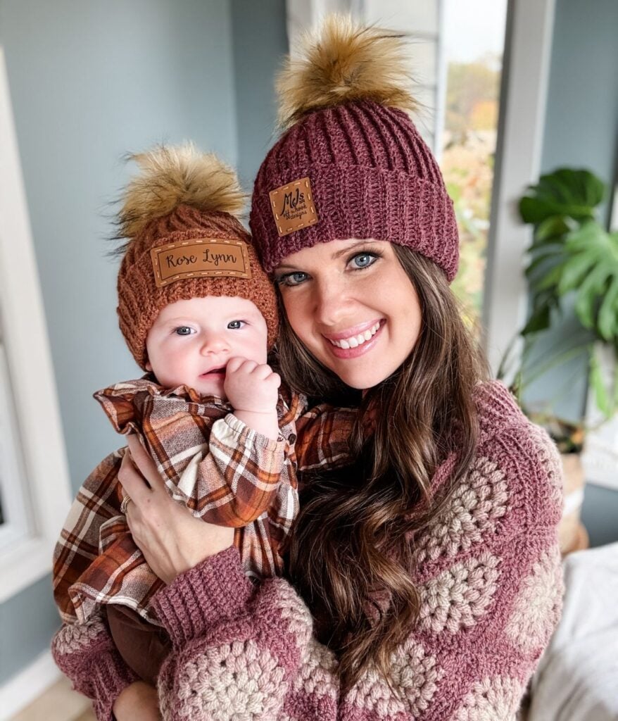 A woman and a baby wearing matching knit hats from MJ's Off The Hook Designs, complete with faux fur poms and coordinated plaid and knit outfits, smiling indoors near a window and a plant.
