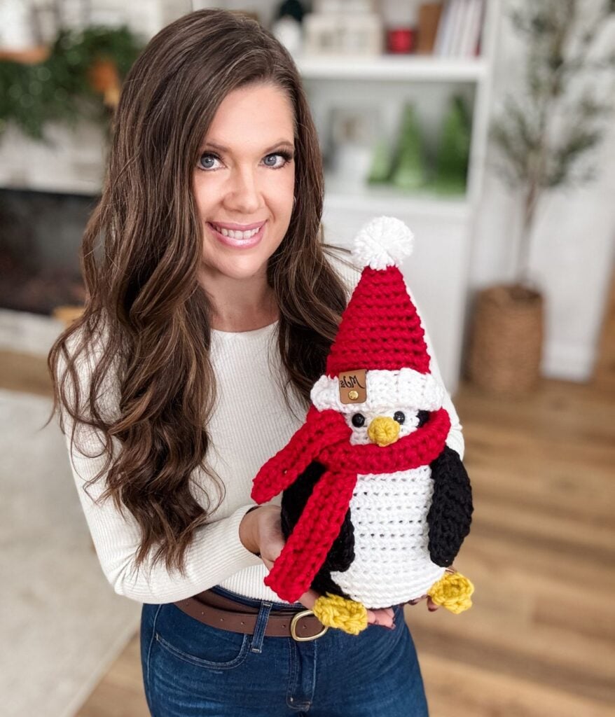 A woman with long brown hair holds a crocheted Santa Penguin wearing a red hat and scarf, standing in a cozy, well-lit room with a plant and bookshelves in the background.