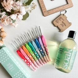 A set of colorful crochet hooks, a bottle of delicate laundry soap, a wooden ruler, a keychain crochet gauge tool, flowers, and a green polka-dot crochet hook box are arranged on a white surface—perfect for any beginner’s guide to crochet.