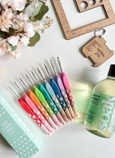A set of colorful crochet hooks, a bottle of delicate laundry soap, a wooden ruler, a keychain crochet gauge tool, flowers, and a green polka-dot crochet hook box are arranged on a white surface—perfect for any beginner’s guide to crochet.
