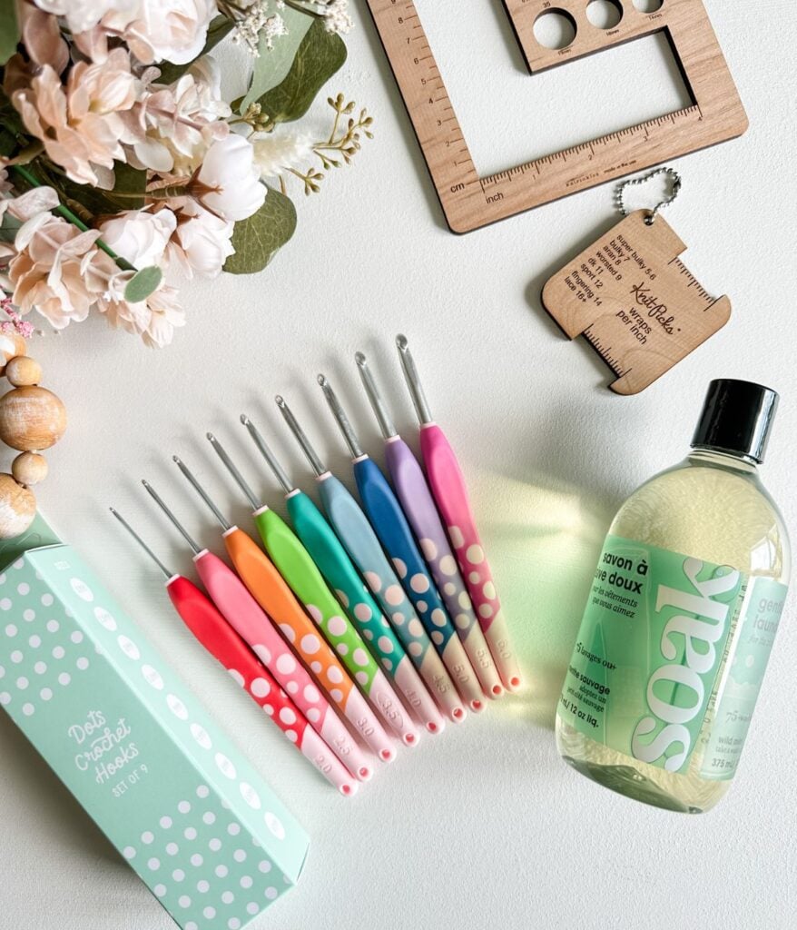 A set of colorful crochet hooks, a bottle of delicate laundry soap, a wooden ruler, a keychain crochet gauge tool, flowers, and a green polka-dot crochet hook box are arranged on a white surface—perfect for any beginner’s guide to crochet.