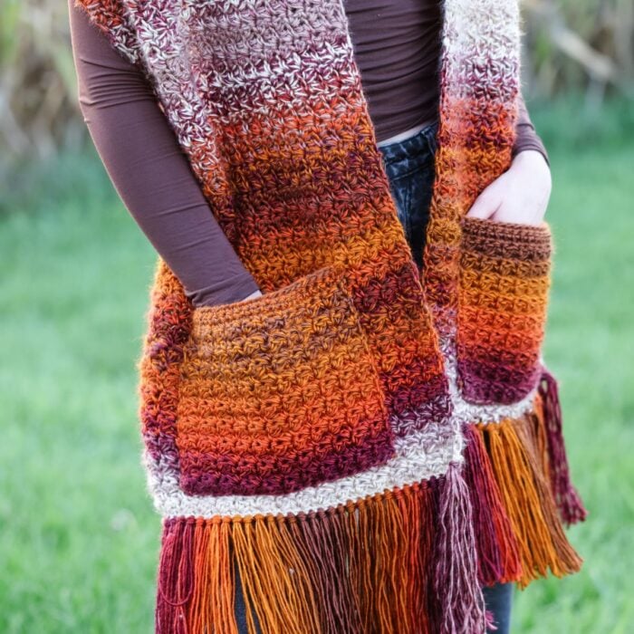 Person wearing a cozy Pocket Shawl with pockets and fringe in orange, red, and brown tones, standing outdoors on grass.
