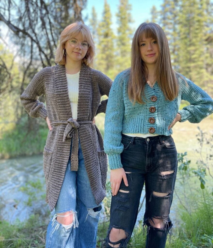 Two young women stand outdoors in front of trees and water, wearing stylish knit cardigans from MJ's Off The Hook Designs paired with ripped jeans.