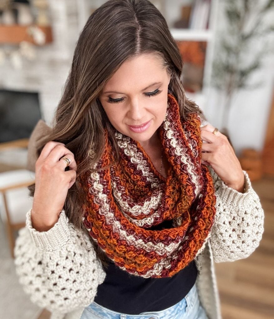 A woman wearing a chunky, multicolored crochet infinity scarf and a cream knit cardigan stands indoors looking down.