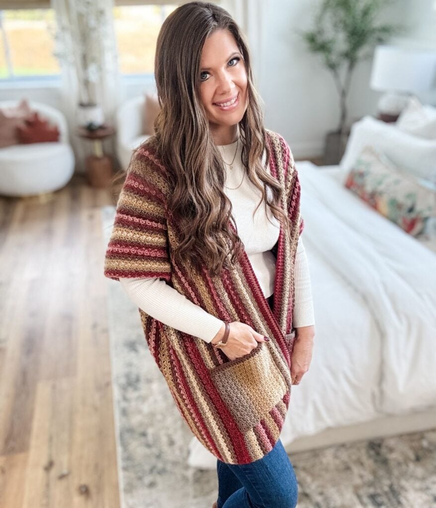 A woman stands in a bedroom, wearing the Suzette Ruana striped knit cardigan over a white top and jeans, smiling at the camera. The room features a bed, rug, plants, and large windows.