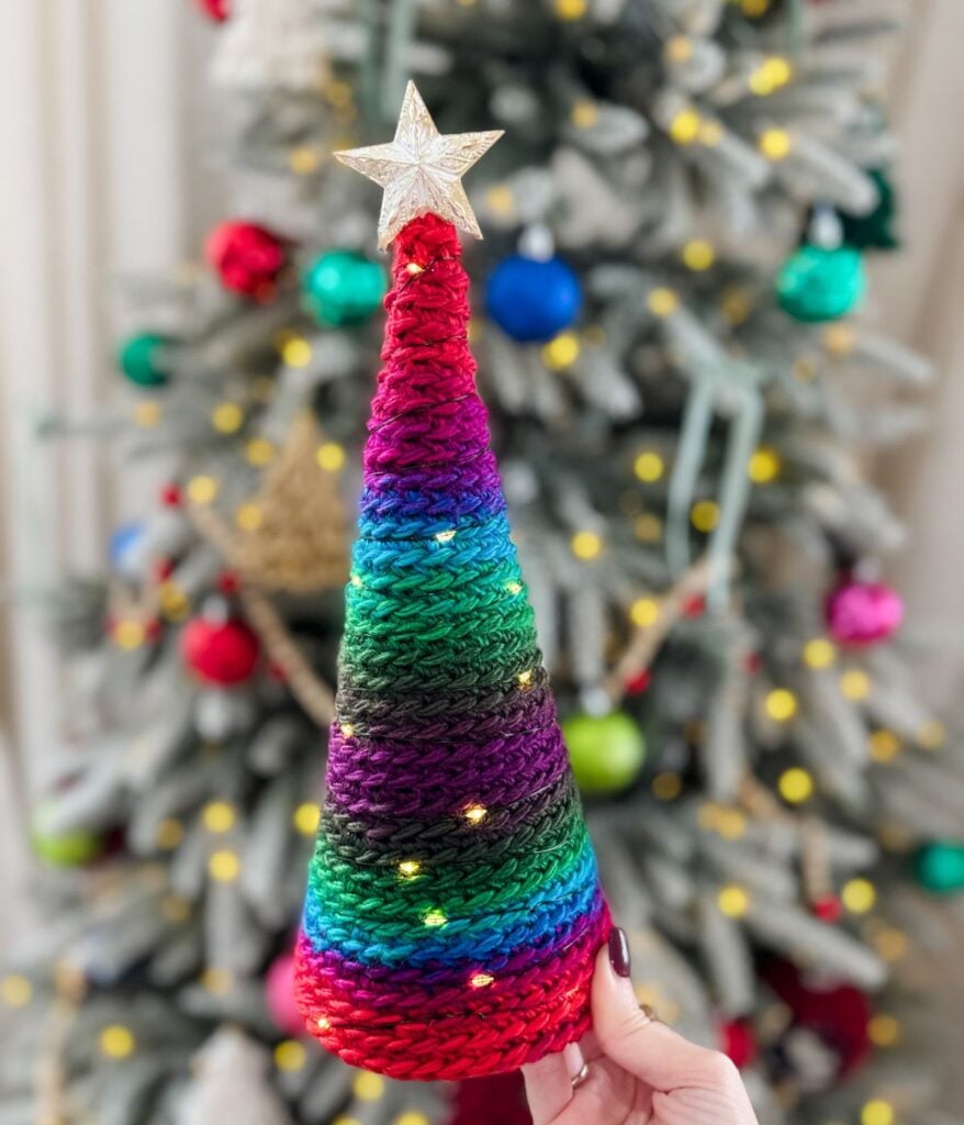 A hand holds a cone-shaped, knitted Christmas decoration with rainbow stripes and a star on top—crafted with the same cozy technique used in a Pocket Shawl—with a decorated Christmas tree in the background.