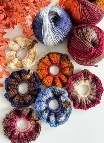 Seven crochet scrunchies in various colors, made with the Suzette stitch, are arranged next to three balls of yarn and orange foliage on a white surface.