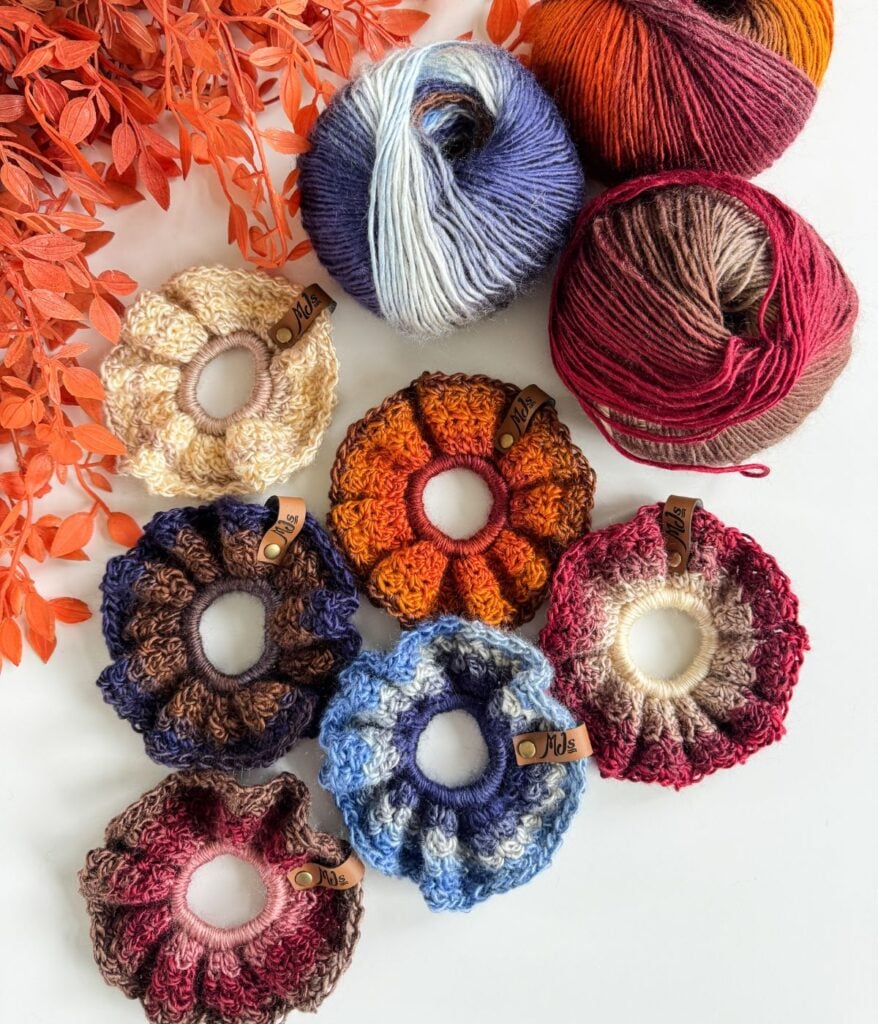 Seven crochet scrunchies in various colors, made with the Suzette stitch, are arranged next to three balls of yarn and orange foliage on a white surface.