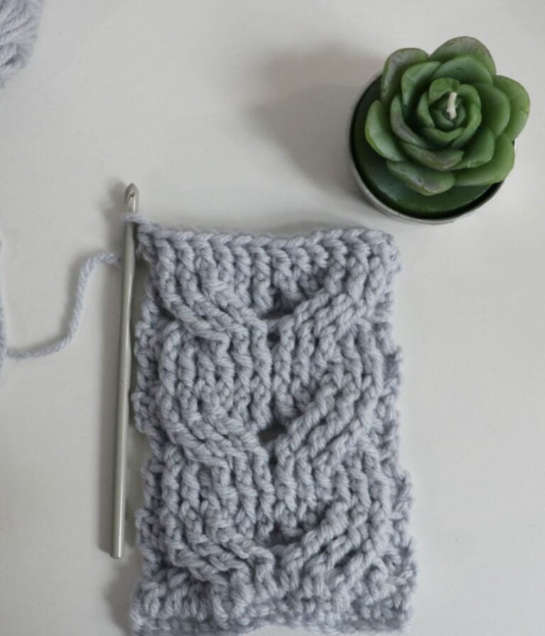 A partially crocheted gray swatch with a textured pattern, a crochet hook, and a small potted succulent on a white surface.