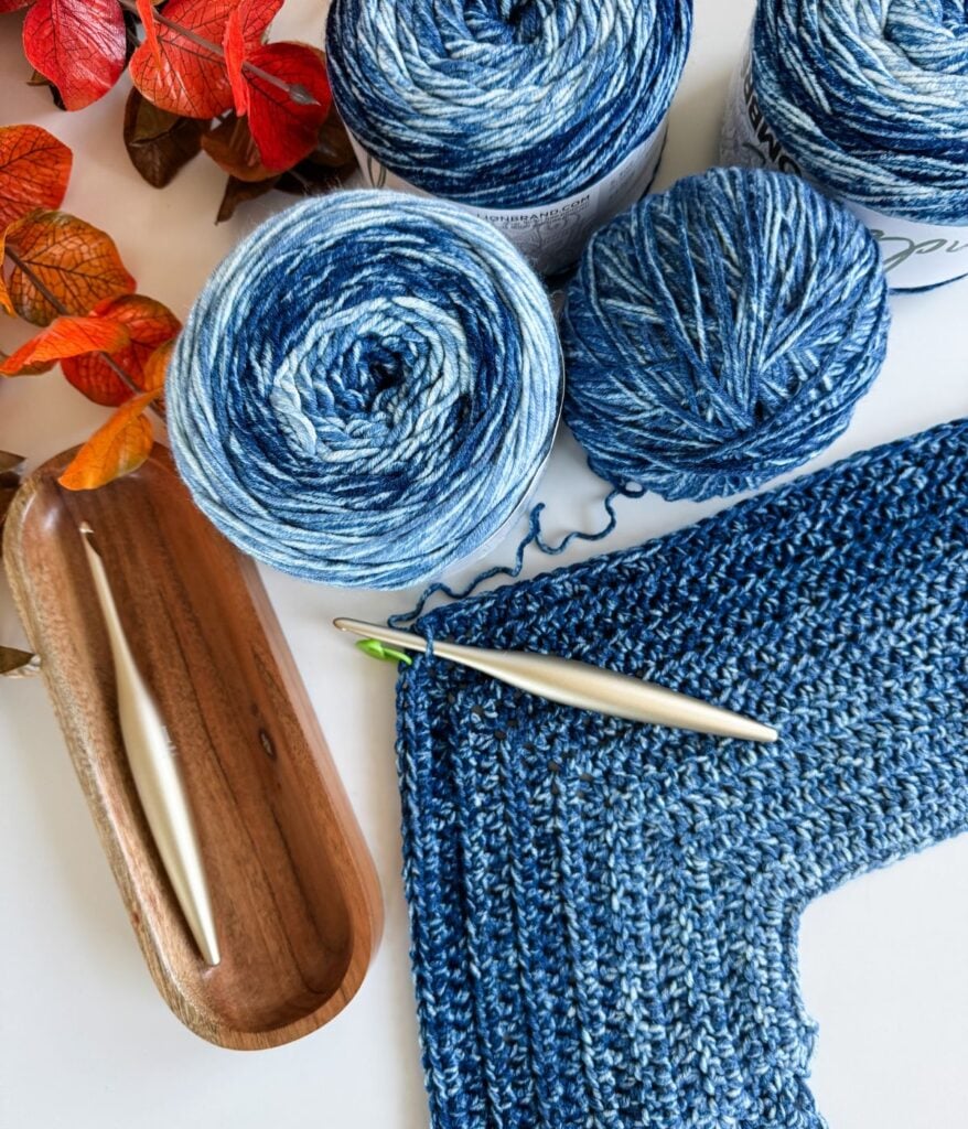 Blue and white yarn balls, a silver crochet hook, a crocheted fabric piece, and a wooden tray with another hook are arranged on a white surface with orange artificial leaves—perfect inspiration for your next everyday crochet pattern.