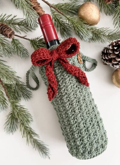 A wine bottle in a green crocheted wine cozy with a red bow, surrounded by pine branches, a pinecone, and gold ornaments on a white surface.