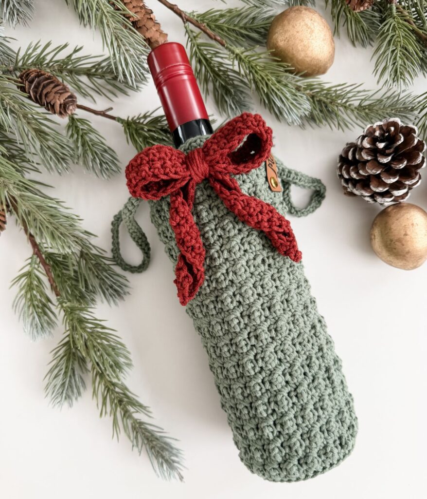 A wine bottle in a green crocheted wine cozy with a red bow, surrounded by pine branches, a pinecone, and gold ornaments on a white surface.