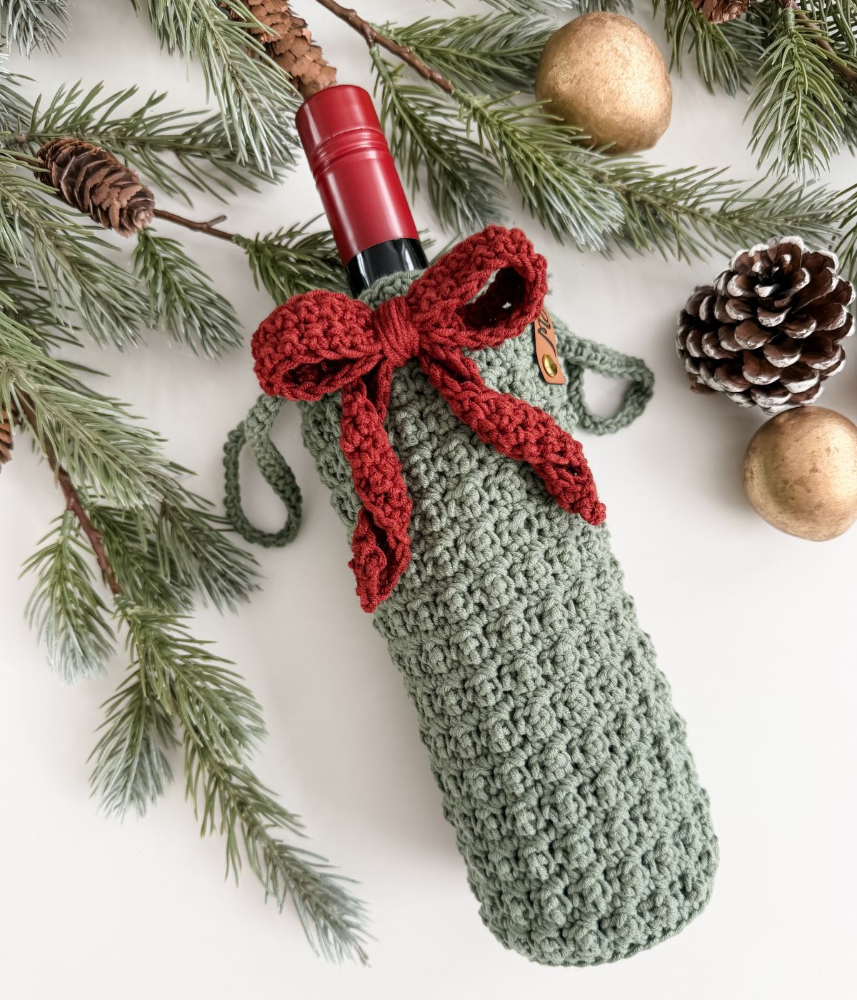 A wine bottle in a green crocheted wine cozy with a red bow, surrounded by pine branches, a pinecone, and gold ornaments on a white surface.