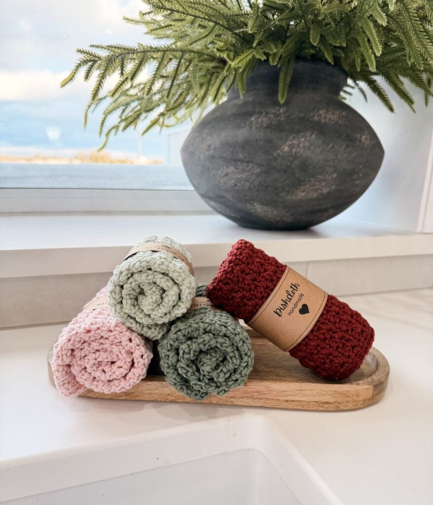 Four rolled knitted dishcloths in pink, green, and red are displayed on a wooden tray in front of a window with a large vase of green fern leaves in the background.