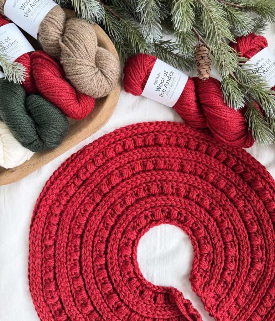 A partially crocheted red tree skirt is displayed near skeins of yarn in red, green, white, and brown, with pine branches and a pinecone above—a cozy scene perfect for crafting a Crochet Snowman.