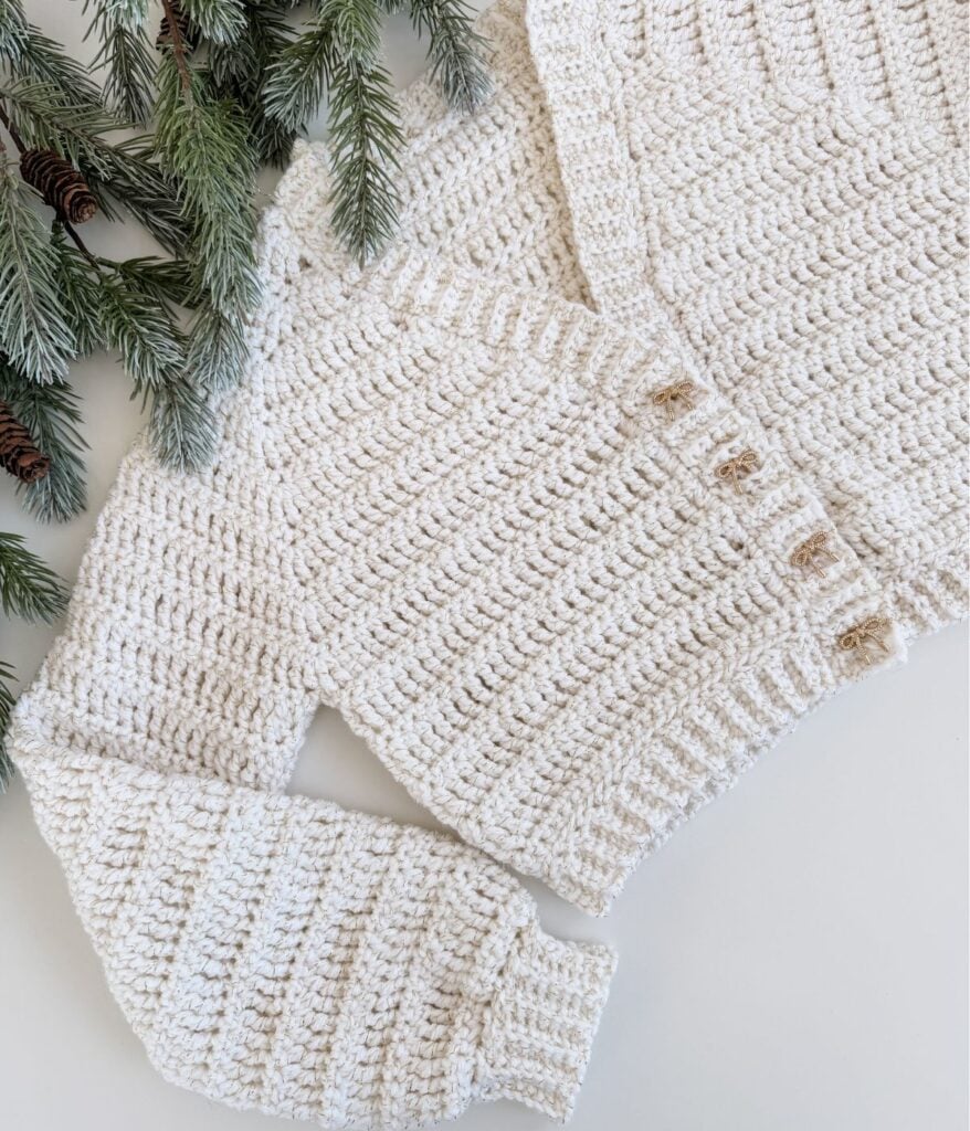 A cream-colored crocheted cardigan with ribbed sleeves and front detailing is laid flat near green pine branches on a white surface.