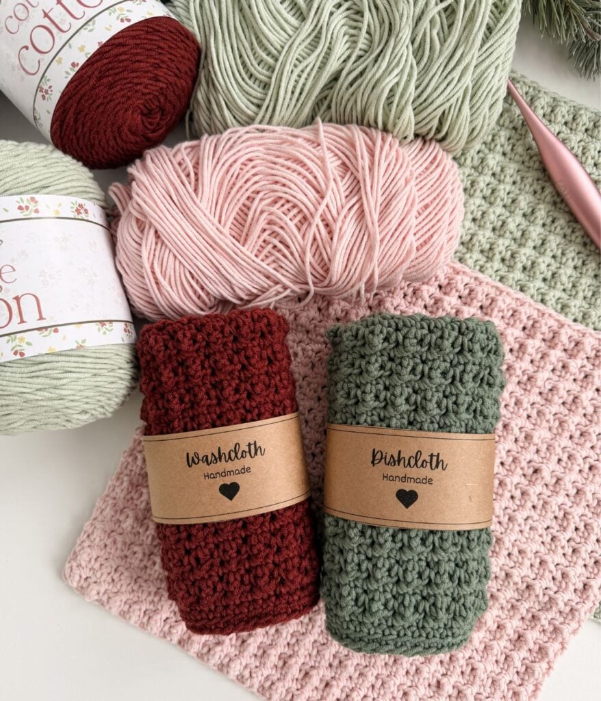 Two handmade crocheted dishcloths, one red and one green, are labeled and placed on top of skeins of yarn and a pink crocheted cloth, with a crochet hook nearby.