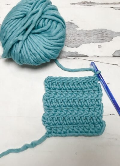 A ball of thick turquoise yarn, a swatch of crocheted fabric, and a transparent blue crochet hook on a white wooden surface.