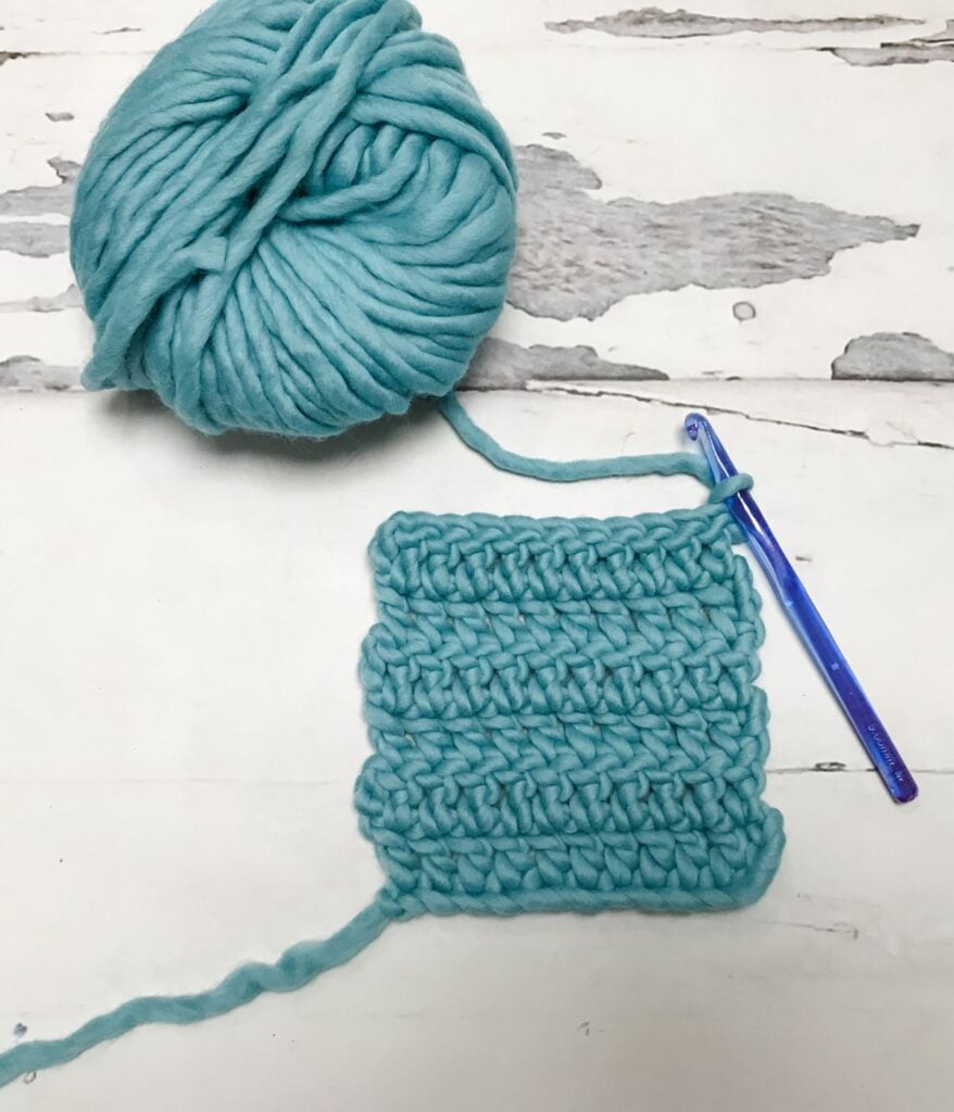 A ball of thick turquoise yarn, a swatch of crocheted fabric, and a transparent blue crochet hook on a white wooden surface.