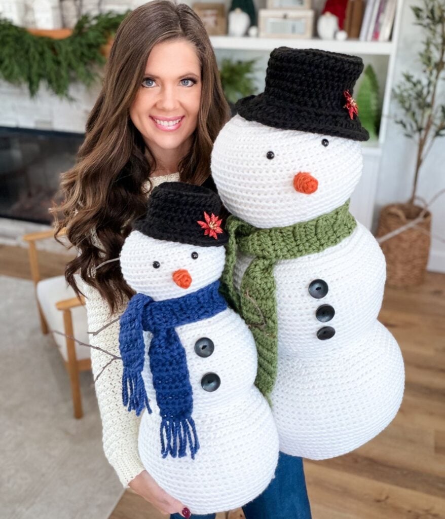 A woman with long brown hair holds two Crochet Snowman figures, one with a blue scarf and the other with a green scarf, in a cozy indoor setting.