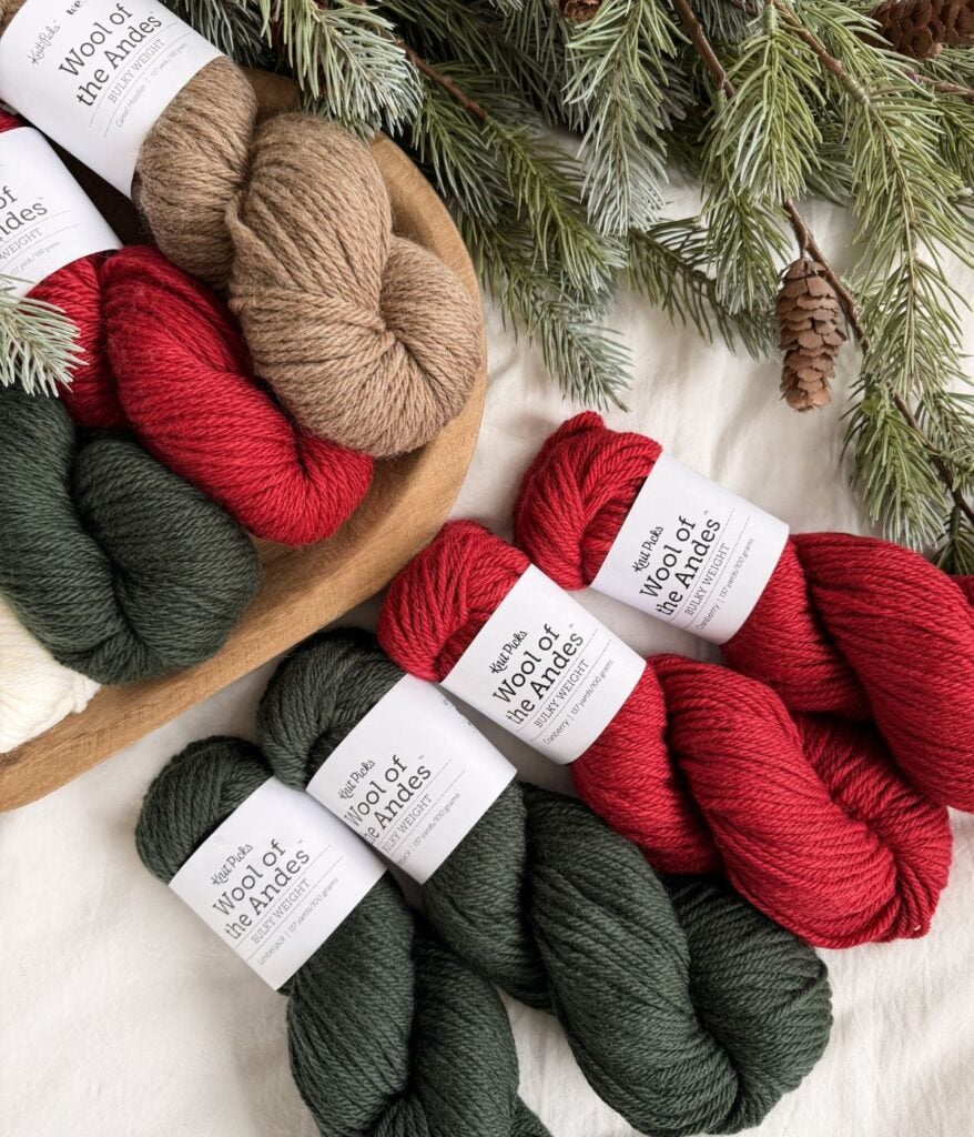 Skeins of red, green, beige, and white yarn labeled "Wool of the Andes" are arranged on a white surface with pine branches and cones nearby—perfect for crafting a Crochet Snowman.