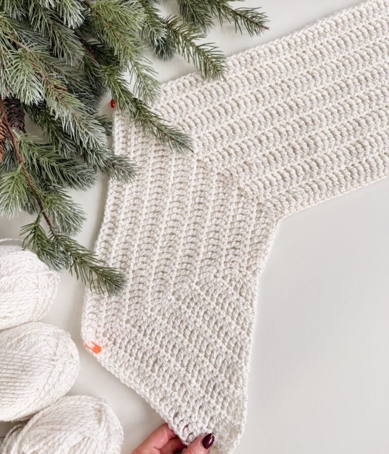 A hand holds the corner of a crocheted white cardigan piece, surrounded by pine branches and three skeins of white yarn on a white surface.