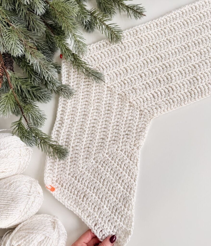 A hand holds the corner of a crocheted white cardigan piece, surrounded by pine branches and three skeins of white yarn on a white surface.