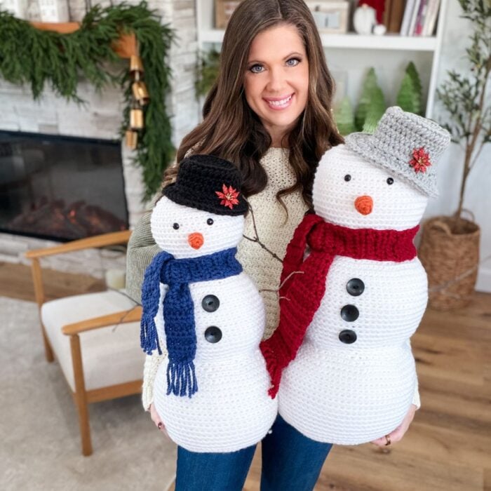 A woman indoors holds two Crochet Snowman figures dressed in scarves and hats, standing in a cozy living room adorned with festive holiday decorations.
