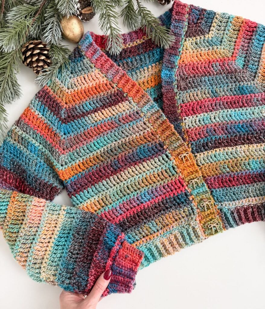 A colorful crocheted cardigan with a chevron pattern is displayed on a white surface, near pine branches, pine cones, and gold ornaments—the perfect festive cardigan for cozy winter days.