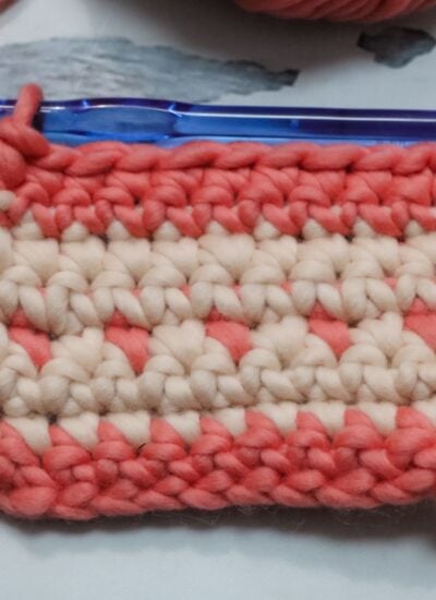 A small swatch of crochet fabric in pink and cream yarn with a blue crochet hook inserted in the top row.
