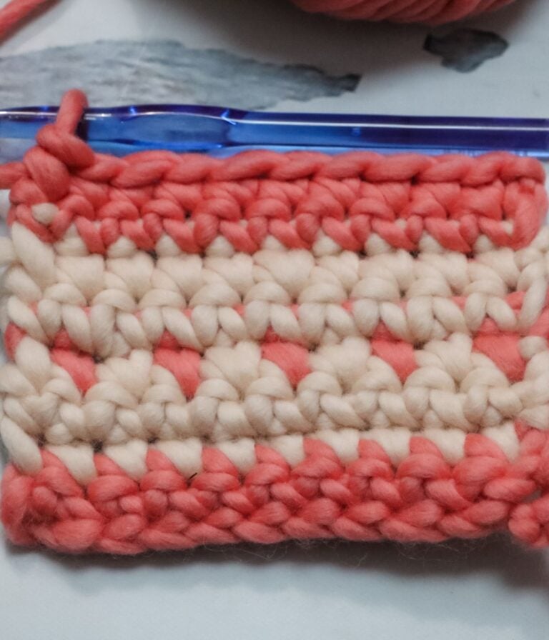 A small swatch of crochet fabric in pink and cream yarn with a blue crochet hook inserted in the top row.