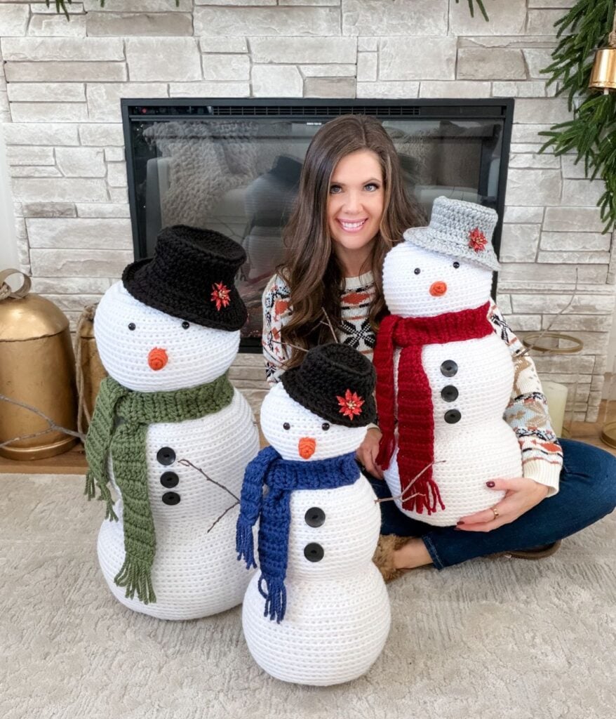 A woman sits on the floor in front of a fireplace, smiling and holding three crochet snowman figures, each adorned with colorful scarves and hats.