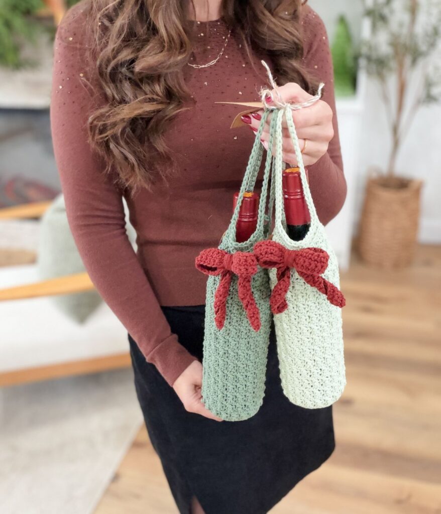 A woman holds two wine bottles in light green crocheted wine cozies with red bows, wearing a brown long-sleeve top and black skirt, indoors.