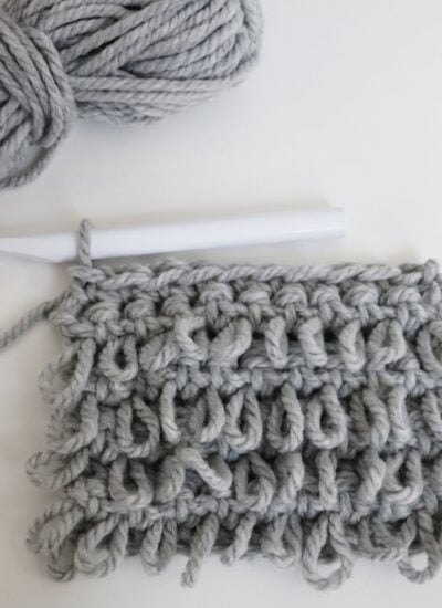 A partially crocheted swatch made with thick, light gray yarn and a white crochet hook resting on a white surface.