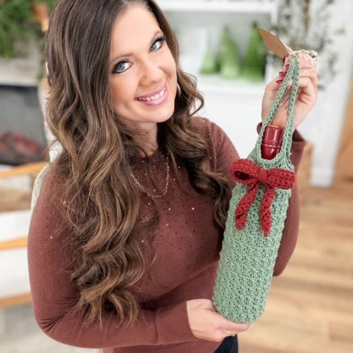 Woman holding a green crocheted wine cozy with a red bow, standing indoors in a cozy living room setting.