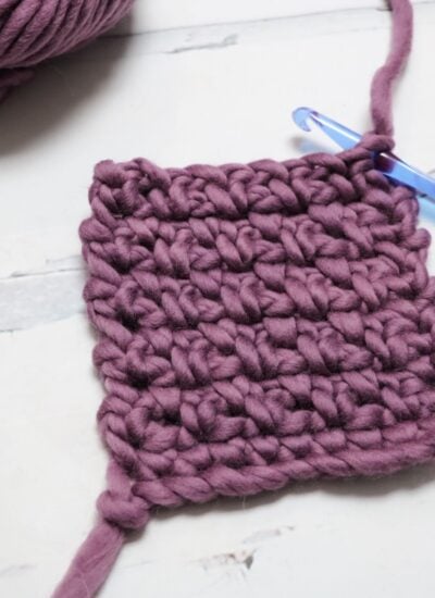 A square swatch of chunky purple crochet yarn with a blue crochet hook attached, resting on a light surface.