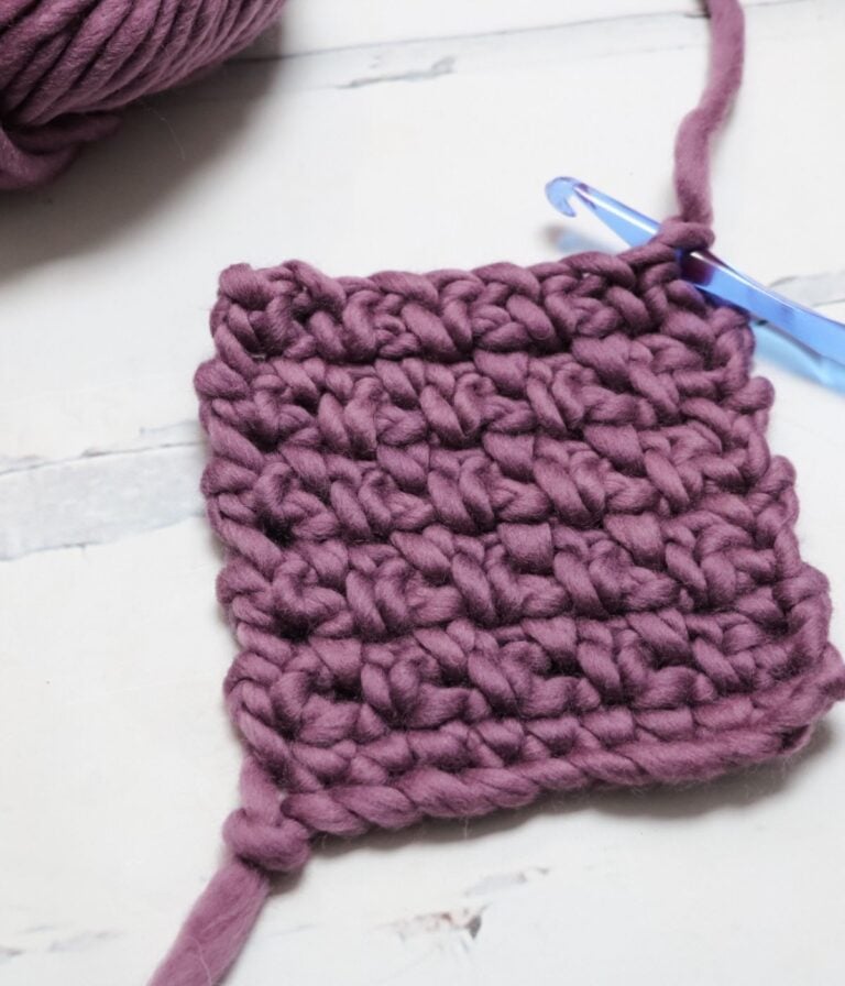 A square swatch of chunky purple crochet yarn with a blue crochet hook attached, resting on a light surface.